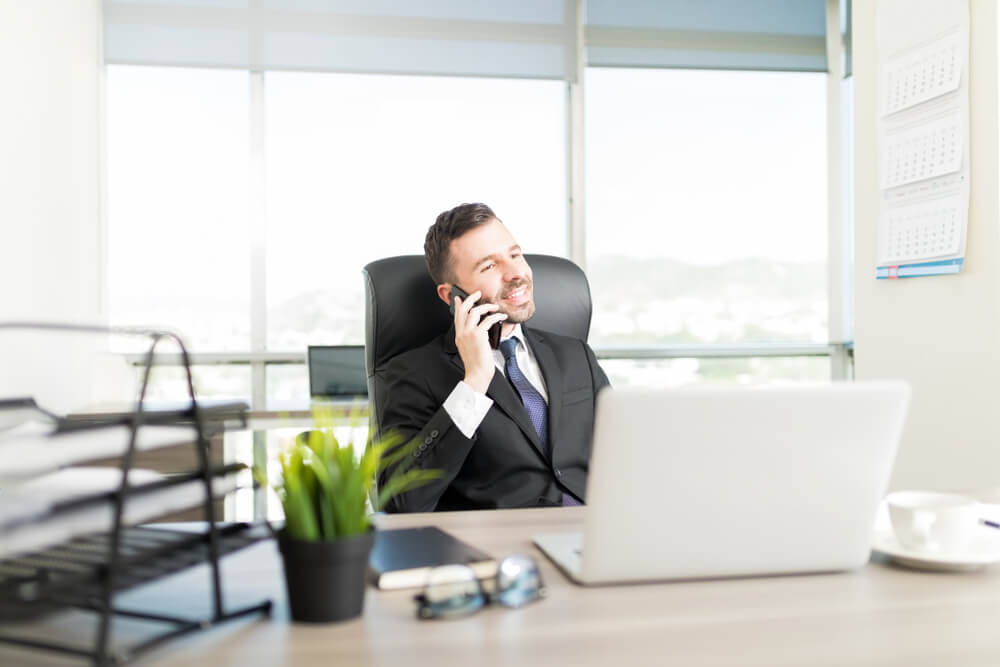 Mid Adult Entrepreneur in Suit Smiling While Having Smartphone Conversation in His Own Office