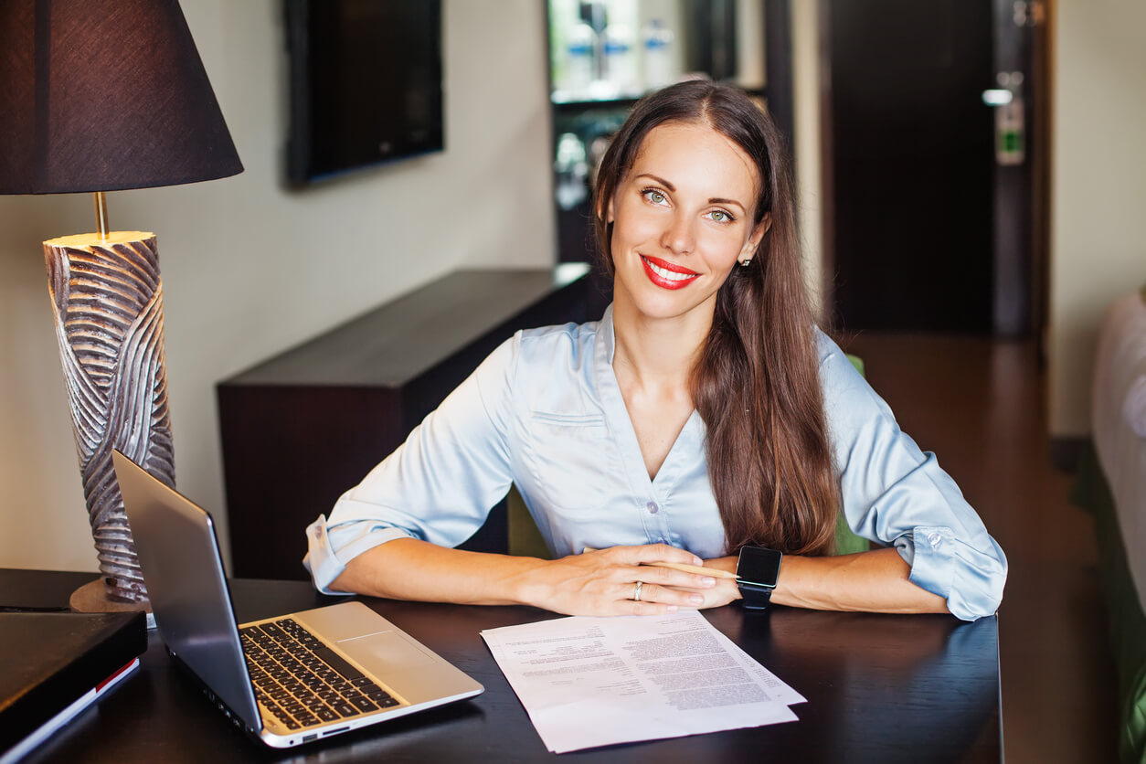 Beautiful Business Woman in Casual Dress Sitting At a Table With Laptop Looking at Camera and Smiling