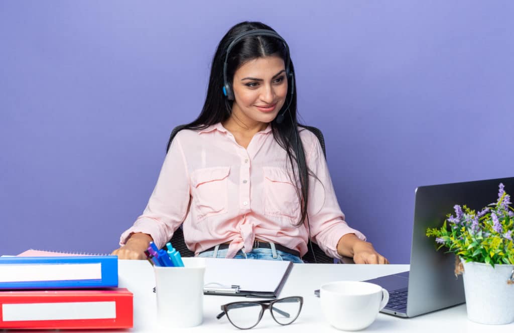 young beautiful woman in casual clothes wearing headset with microphone sitting at the table with laptop looking at screen smiling confident over blue background working in office